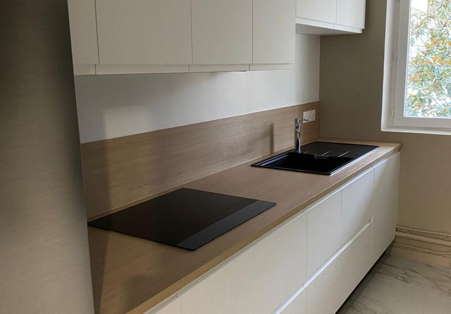 Cuisine linéaire moderne avec meubles blancs sans poignée, plan de travail et crédence en bois clair, évier noir et sol effet marbre.