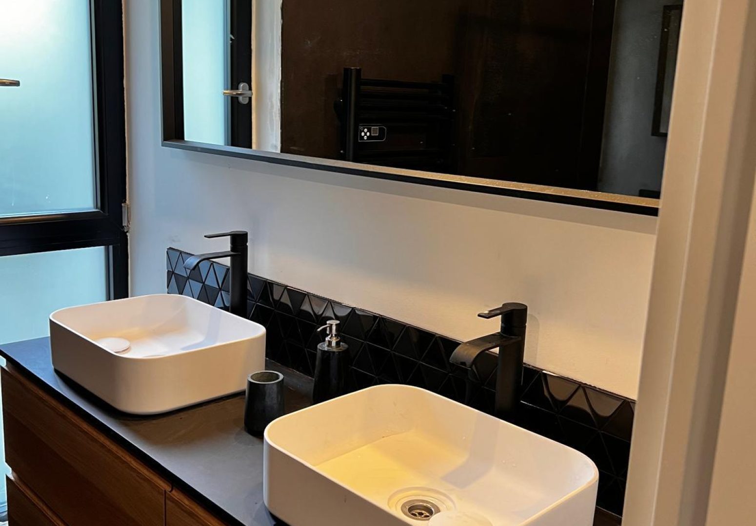 Salle de bain contemporaine avec grand meuble double vasque en bois, robinetterie noire, crédence en faïence géométrique et grand miroir.