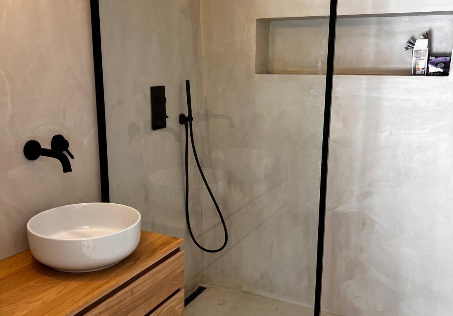 Salle de bain moderne en béton ciré beige avec douche à l'italienne, paroi à profilé noir mat, meuble vasque en bois suspendu et robinetterie noire.